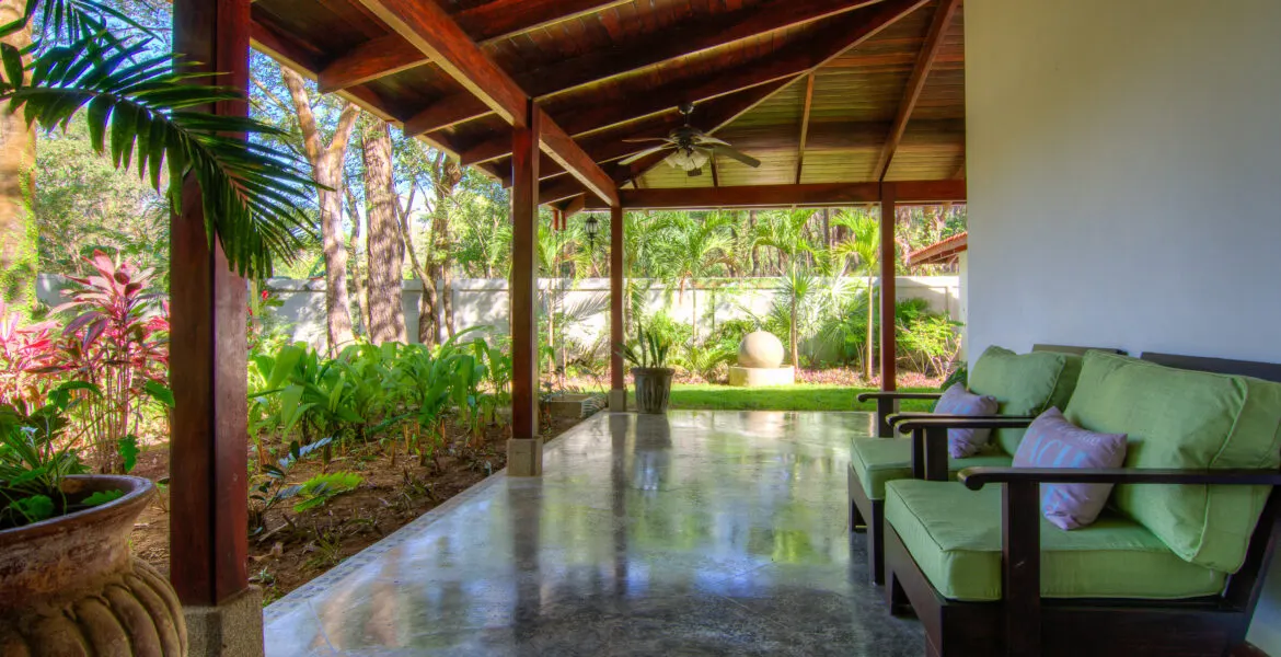 A covered open-air terrace with a dark timber beam ceiling and ceiling fan, two lime-green cushioned armchairs at right, and the lawn and a stone sphere sculpture beyond.