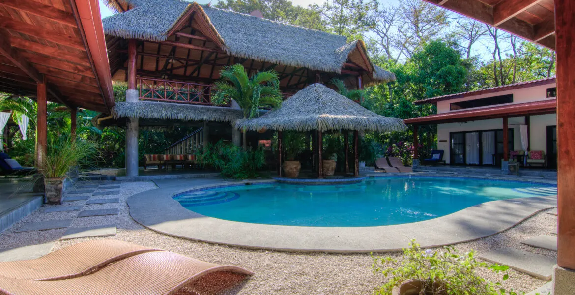 The pool courtyard seen from under a terrace roof: the curved saltwater pool, a freestanding thatched palapa bar at centre, the two-storey thatched main building behind and rattan loungers in the foreground.