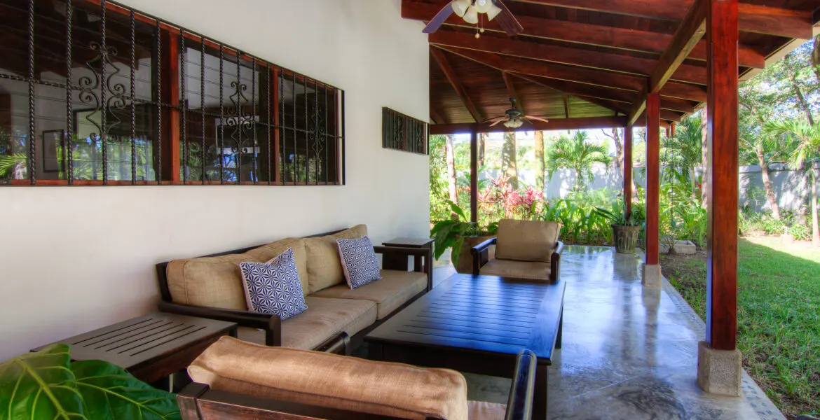 A covered lounge with a tan sofa set and blue geometric cushions around a dark wood coffee table, a decorative iron-grille window wall at left and greenery and lawn at right.