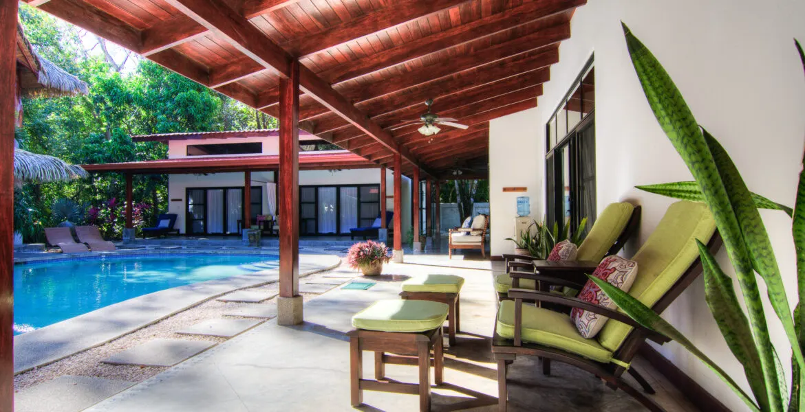 A poolside terrace under a red-stained beam roof with green lounge chairs and ottomans, the pool cutting in from the left and a guest-room building with white curtains across the water.