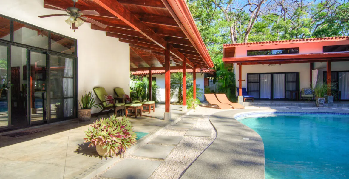 The pool edge with a pebbled deck and stepping-stone path, glass sliding doors at left, orange sun loungers and the red-roofed guest building at right.