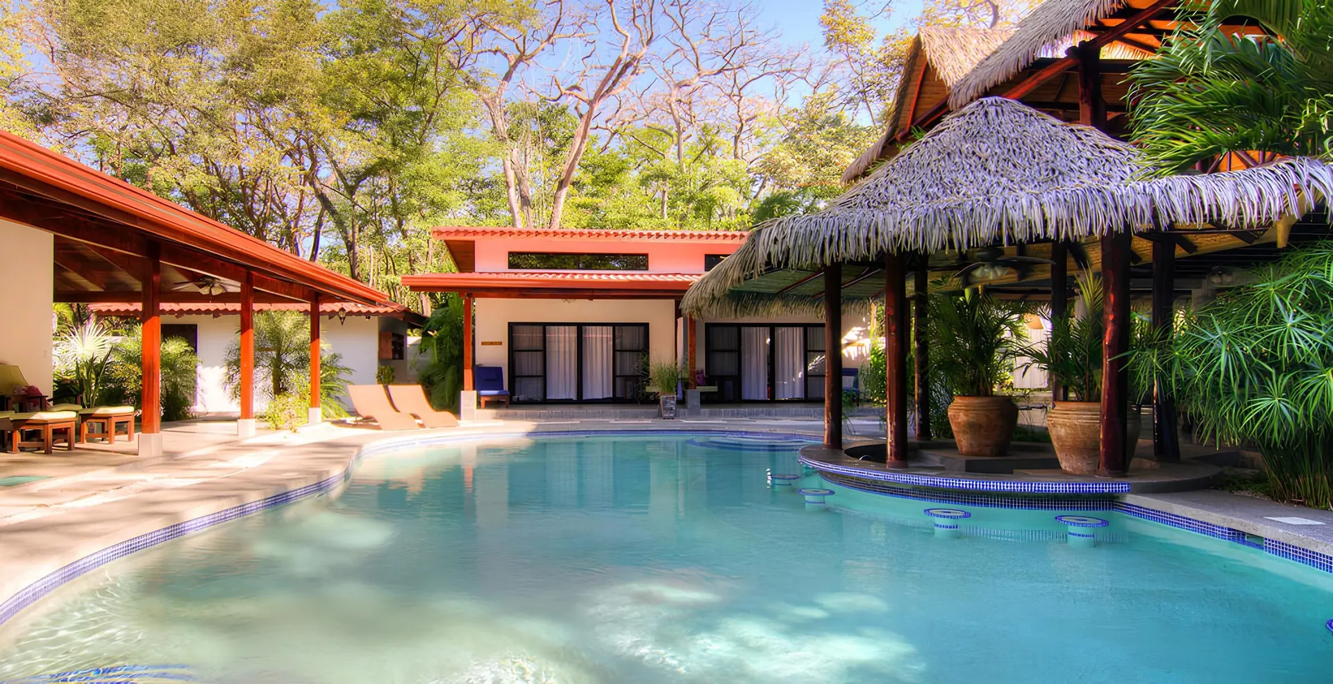 A turquoise saltwater pool filling the foreground, a row of submerged bar stools along a swim-up bar under a large thatched palapa at right, and the red-roofed bedroom wing across the water.