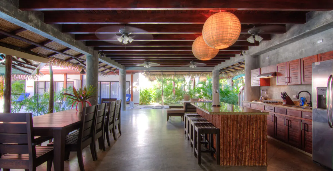 The open great room shot down its long axis: a dark-wood dining table and bench at left, a bamboo-clad kitchen island with bar stools at right, timber beams and woven globe pendants receding toward a bright green opening.