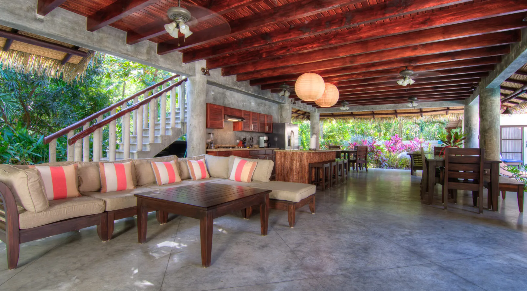 The open ground-floor pavilion: a large sectional sofa with coral-striped cushions, concrete stairs with a rustic log balustrade at left, a bamboo-clad kitchen island mid-frame and a long dining table at right, under an exposed timber and thatch roof.