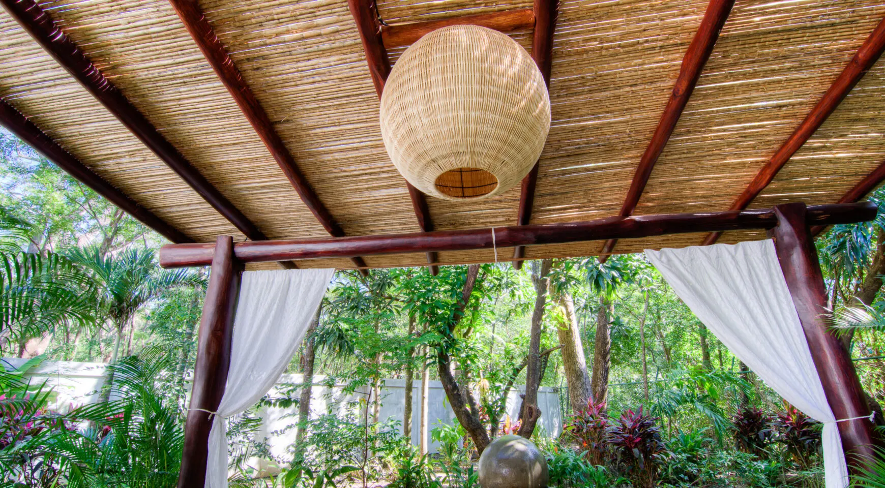 The inside of the meditation pavilion looking out: a bamboo ceiling, a large woven globe lantern, rustic timber framing, white gauze curtains, and a stone sphere in the garden beyond.