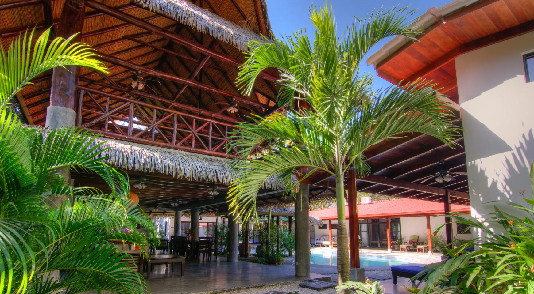The compound courtyard: a soaring thatched roof with exposed timber trusses on the left, a royal palm rising through the centre of the frame, and a red-tiled guest wing beside the pool at right under a deep blue sky.