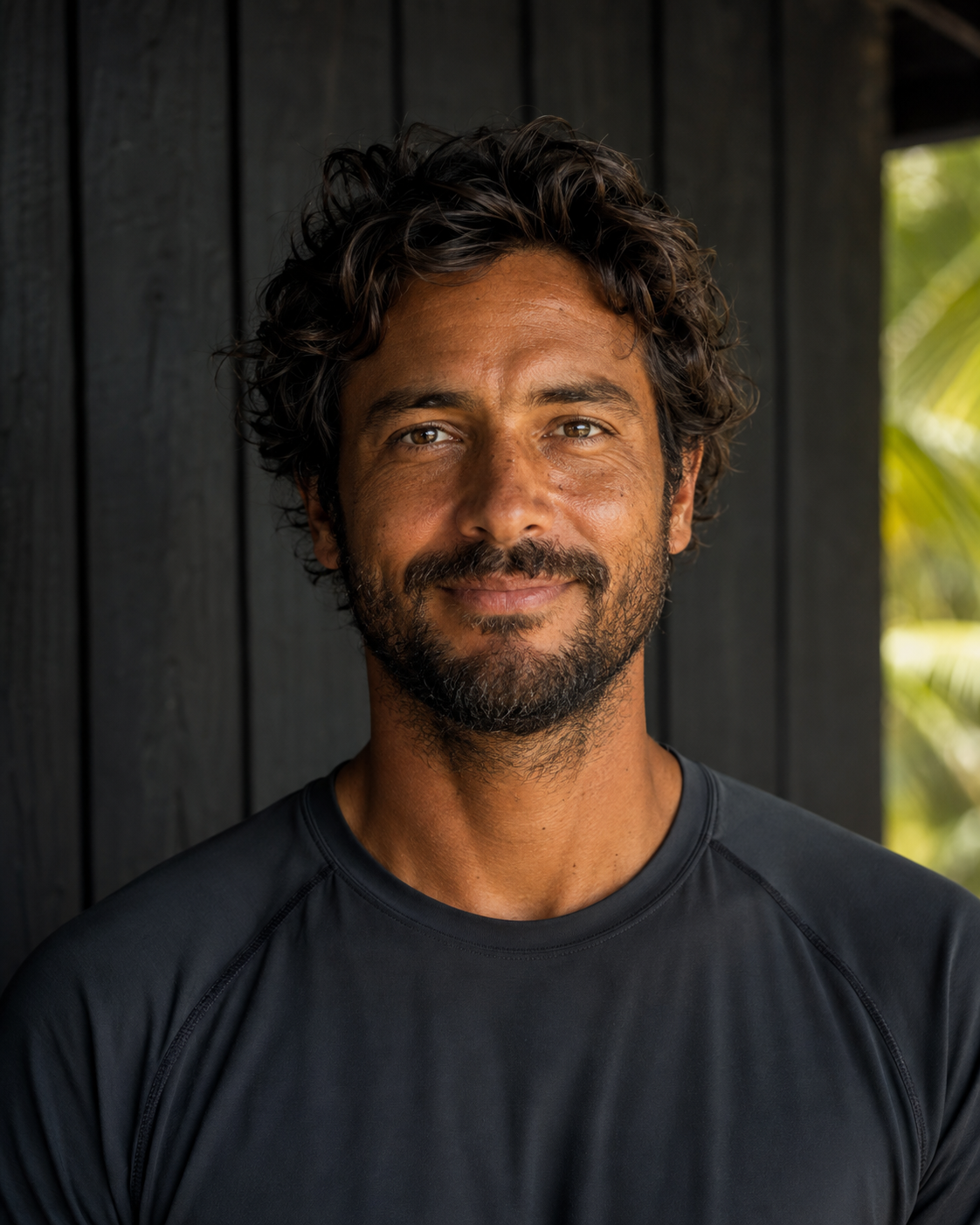 Portrait of Mateo Solís, a SurfWERX guide, in a charcoal technical surf top against a charred-wood wall with tropical foliage behind.