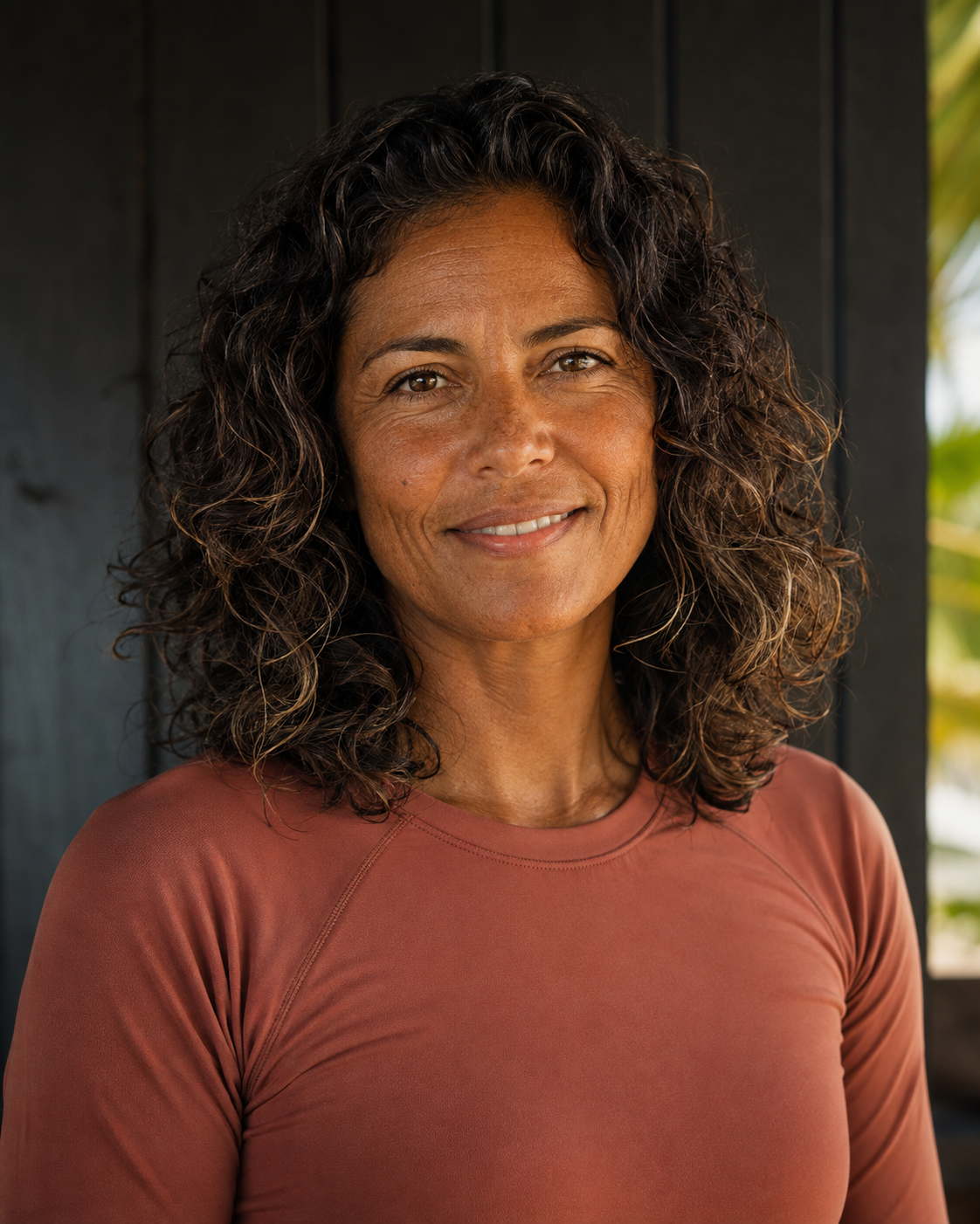 Portrait of Carolina Vargas, a SurfWERX guide, in a terracotta technical surf top against a charred-wood wall with tropical light behind.